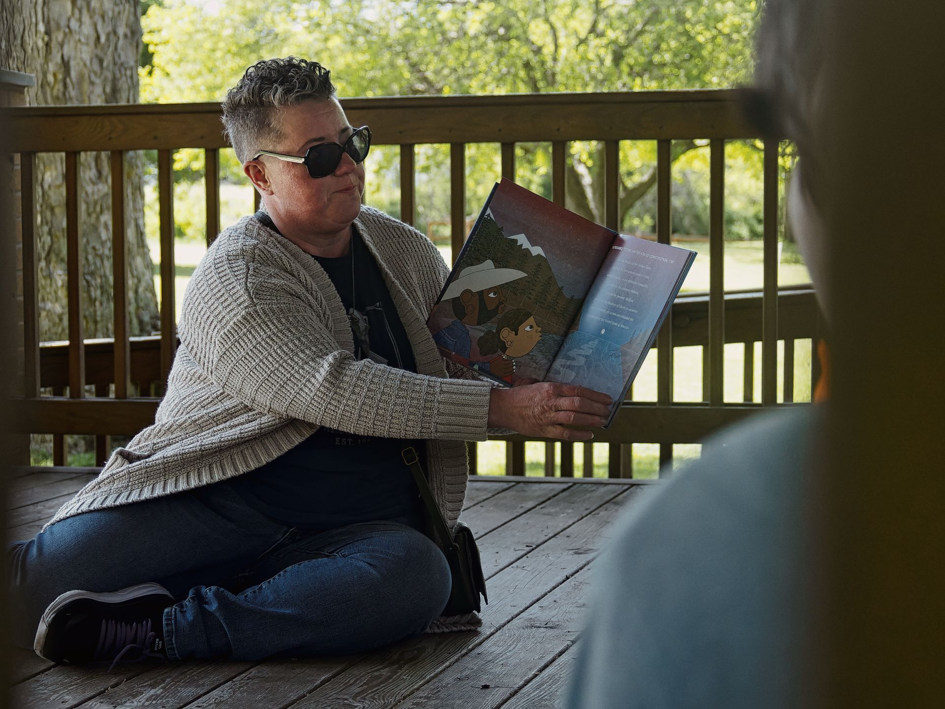 Woman with short salt-and-pepper hair and dark sunglasses sitting cross-legged on a wooden deck, holding open an illustrated children's book toward an unseen listener blurred in the right foreground. The book's spread shows two dark-skinned cartoon children in a moody, deep-blue and red illustrated scene. Green trees and a wood-railed deck are softly blurred in the background.