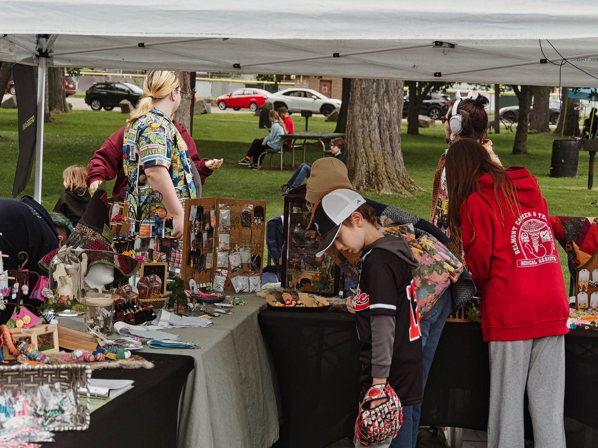 Outdoor craft fair booth beneath a white canopy tent, with tables draped in dark grey cloth and covered in handmade jewelry displays, small figurines, and colorful trinkets. A young boy in a dark baseball jersey and tan cap examines the goods in the foreground, while a woman in a red "Belmont Career & Technical Medical Assistant" hoodie browses to the right; a vendor in a colorful patterned top stands behind the table on the left. A grassy park with parked cars and large tree trunks stretches into the background.
