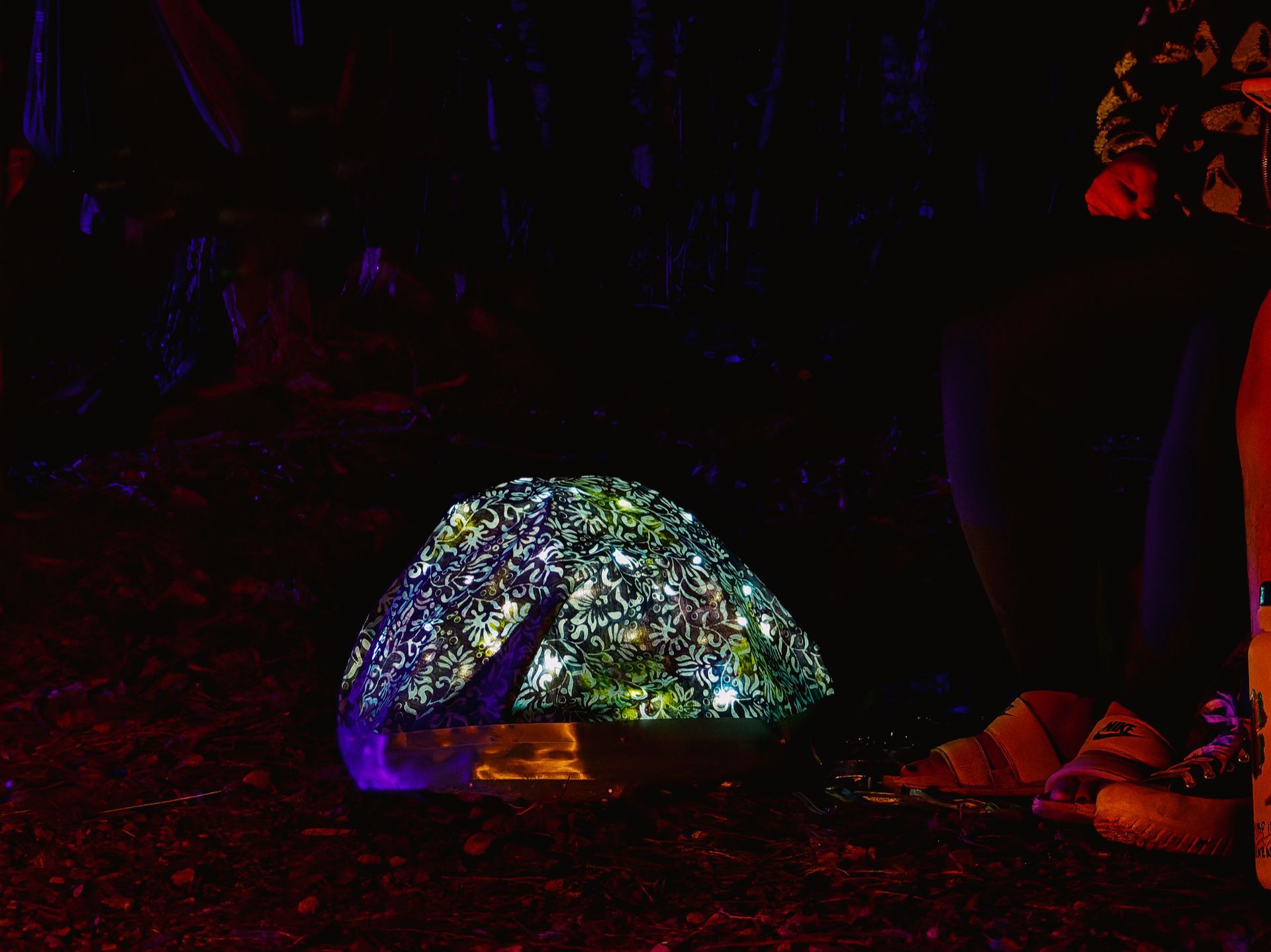 A dome tent covered in an intricate floral and swirling pattern glows from within against a near-total darkness, its fabric illuminated in blues, greens, and whites like a bioluminescent creature. A pair of Nike sandals and scattered gear sit in the foreground to the right on dark gravel, with shadowy tree trunks barely visible behind.