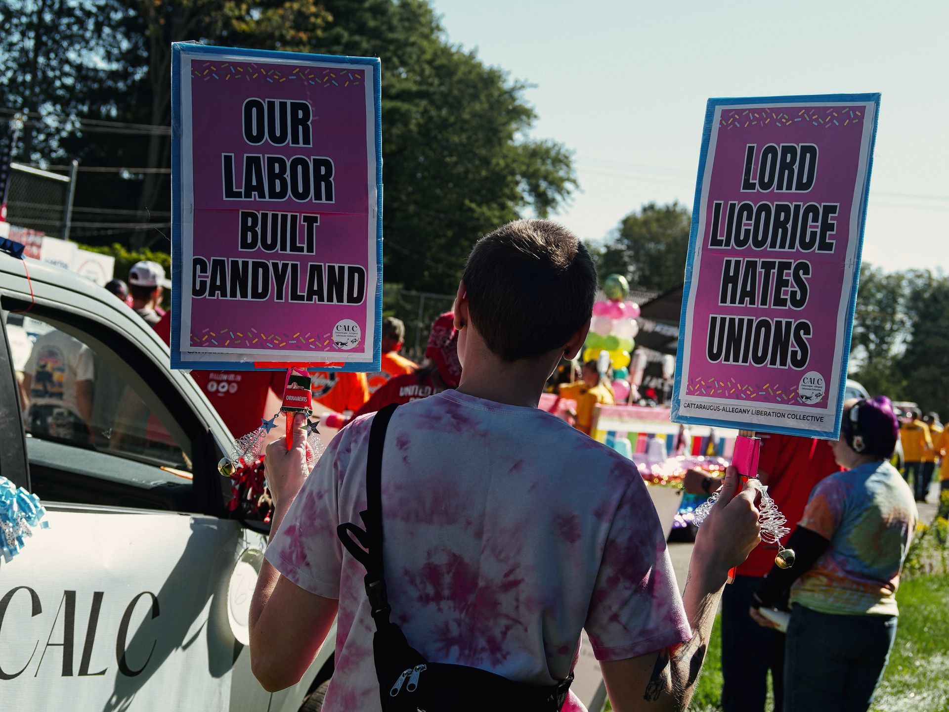 Person in a pink tie-dye shirt, seen from behind, holding two bright magenta protest signs with bold black text — "OUR LABOR BUILT CANDYLAND" on the left and "LORD LICORICE HATES UNIONS" on the right, both branded with the Cattaraugus-Allegany Liberation Collective logo. A colorful parade float and crowd fill the blurred background, with a white vehicle marked "CALC" visible on the left.
