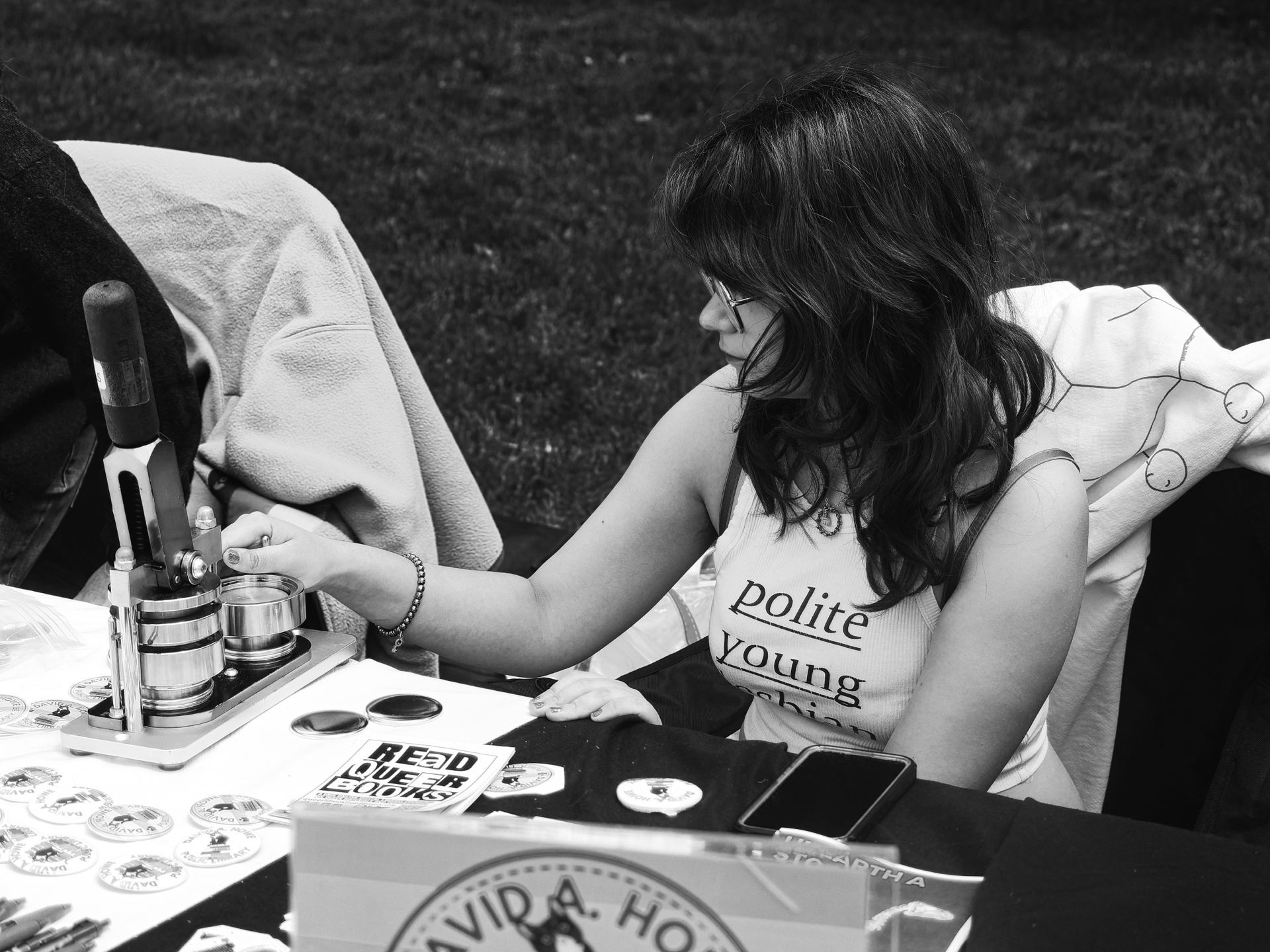 Black-and-white shot of a young woman with long dark hair and glasses leaning over an outdoor table, operating a button-making press. Her shirt reads "polite young" with the bottom text obscured. The table is covered in finished pinback buttons, a sticker reading "READ QUEER BOOKS," and a partially visible sign reading "DAVID A. HO—" in the foreground; grass is visible in the blurred background.