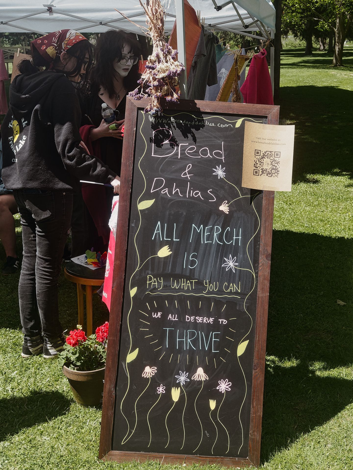 A tall chalkboard sign in a wooden frame reads "Bread & Dahlias / ALL MERCH IS / PAY WHAT YOU CAN / we all deserve to THRIVE," decorated with hand-drawn flowers and leaves, with a QR code taped to the upper right corner. A potted red geranium sits at the base, and two people in dark clothing browse under a white canopy tent to the left, with green grass and trees visible in the sunny background.