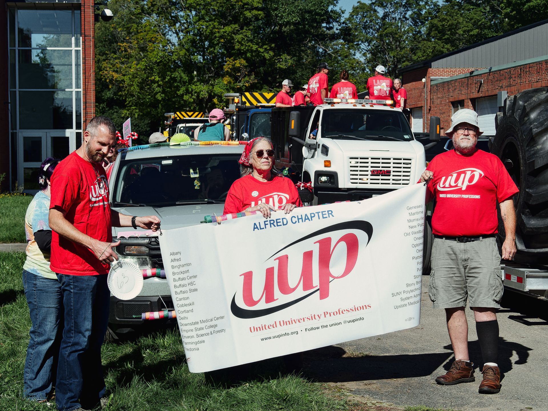 Three people in red UUP shirts hold a white banner reading "ALFRED CHAPTER / uup / United University Professions / www.uupinfo.org • Follow the union @uupinfo" in a parade setting, with a decorated car and a large white truck behind them carrying more red-shirted participants. Trees and brick buildings frame the background under a bright sky.