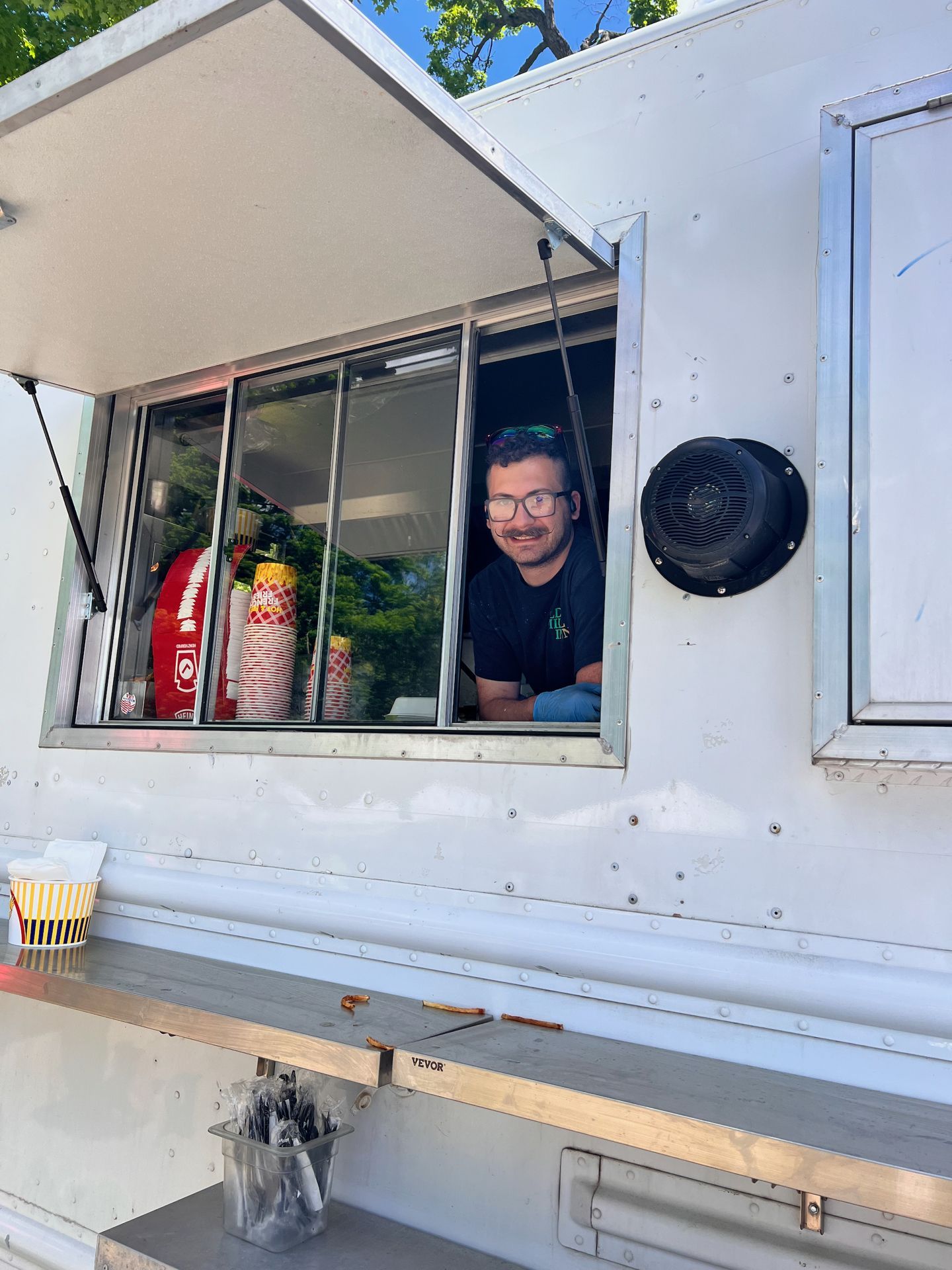 A smiling man with glasses and a mustache leans out of the service window of a white food truck, with stacks of paper cups and condiment bottles visible behind him. A yellow-striped cup sits on the left of the stainless steel counter below, alongside a small caddy of utensils stamped "VEVOR."
