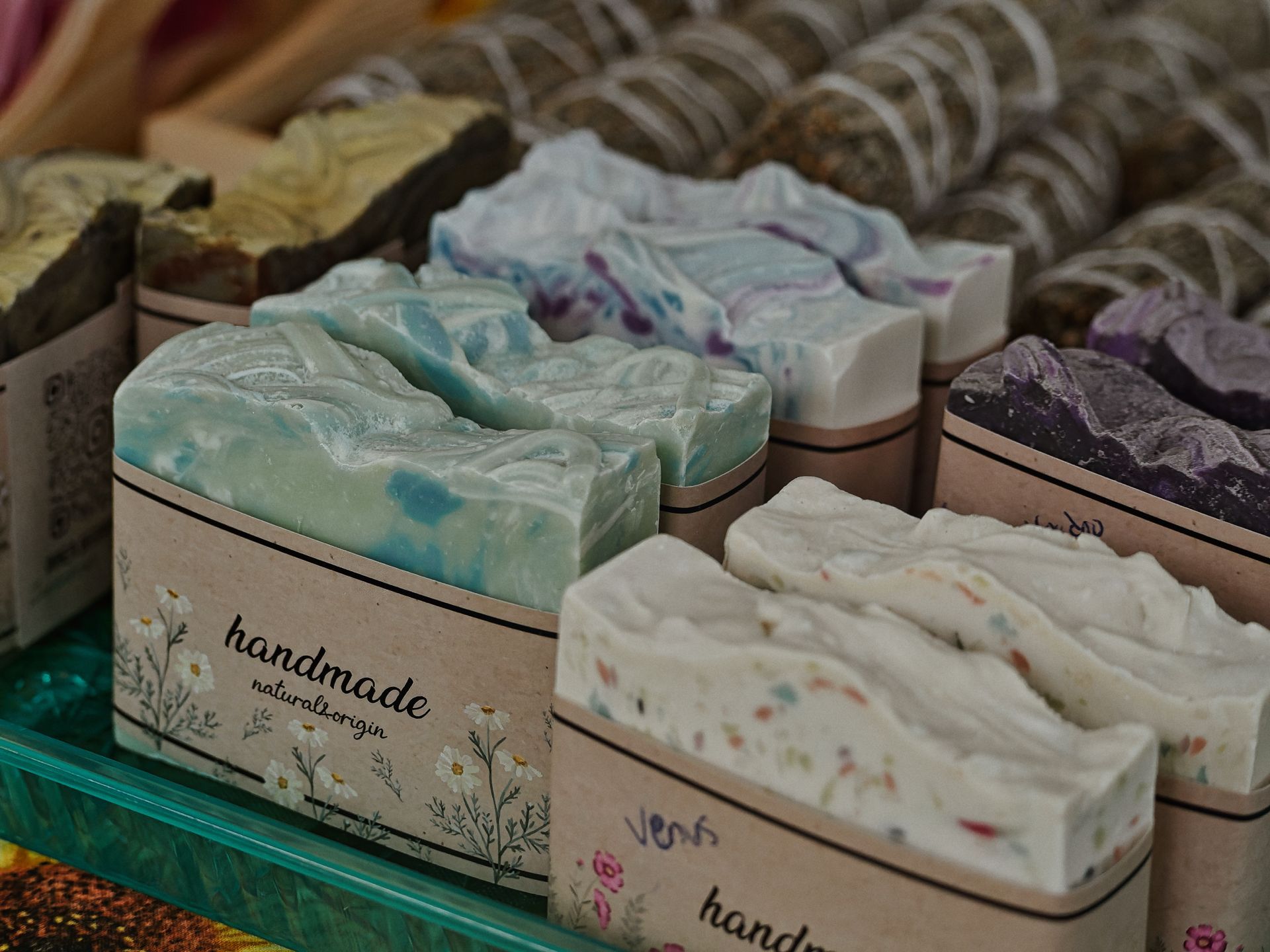 Rows of artisan soap bars displayed on a teal tray, each wrapped in kraft paper bands printed with floral illustrations and the text "handmade natural origin." The bars vary in color — pale mint-blue with swirls in the foreground, white speckled with pastel confetti dots labeled "Venus" in front right, blue-purple swirled and deep purple in the middle row — with additional soaps and bundled herbs blurred in the shallow background.