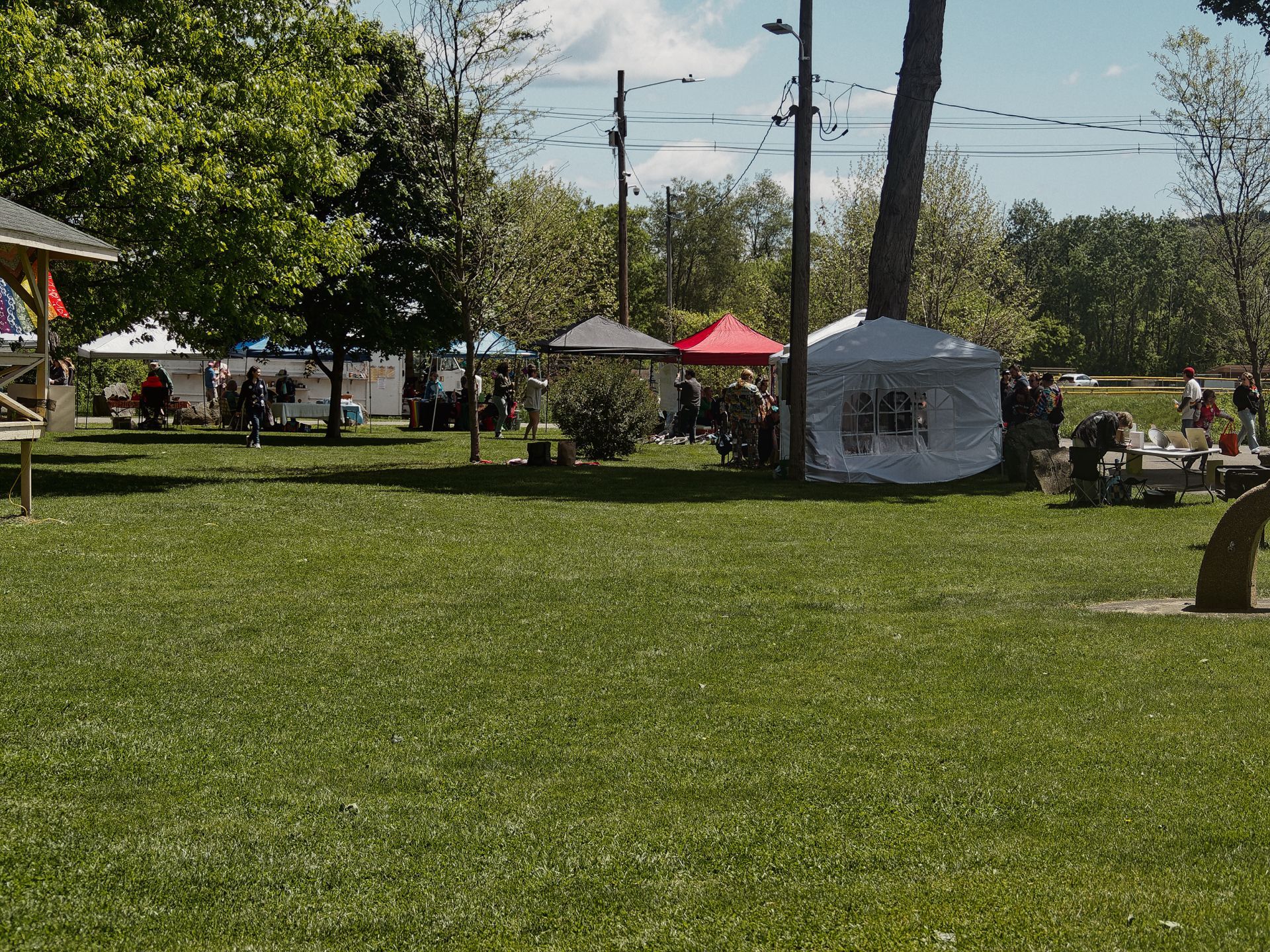 An outdoor community event spread across a wide green lawn, with several vendor canopies in the middle distance — a white gazebo-style tent, a red pop-up, a dark gray canopy, and a white stretch tent to the left — scattered beneath leafy spring trees and utility poles strung with wires. Small groups of people mill between the booths under a partly cloudy sky, with a densely wooded hillside visible in the background.