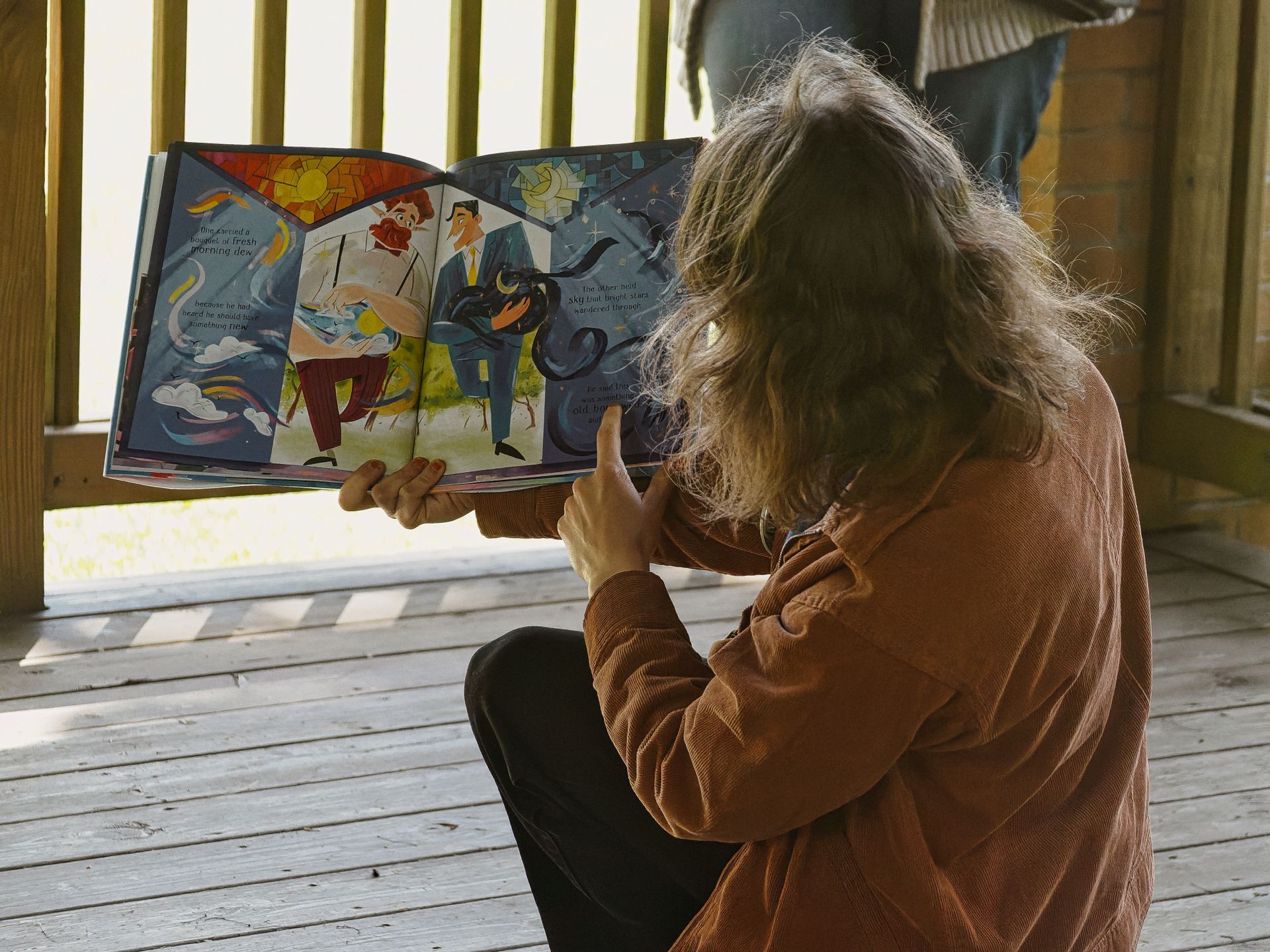 Child with long, windswept hair sitting cross-legged on a weathered wooden porch, holding up an open illustrated picture book. The visible spread shows two painted characters — one kneeling in red pants, one standing in a blue suit — surrounded by swirling dark shapes and stained-glass-style skies, with small blocks of text reading "like a cold a pocket of fresh morning dew" and "The Miser held sky but longed after wanderer thereof."