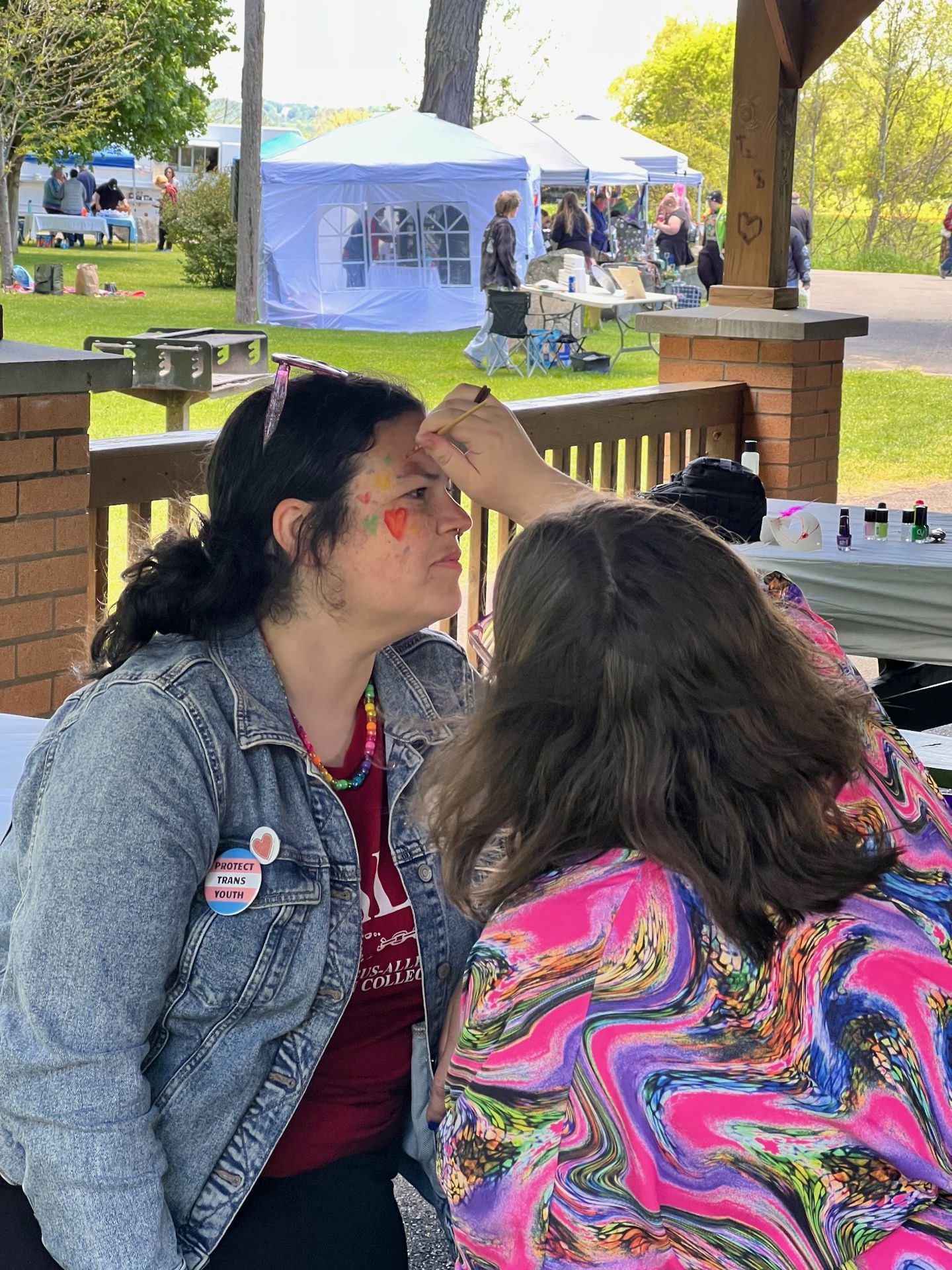 Woman in a denim jacket wearing a "Protect Trans Youth" button and a colorful beaded necklace getting her face painted — a red heart and rainbow dots already on her cheek — by a person in a vibrant pink-and-purple marbled top at an outdoor community event, with white canopy tents, green lawn, and scattered attendees visible in the background.