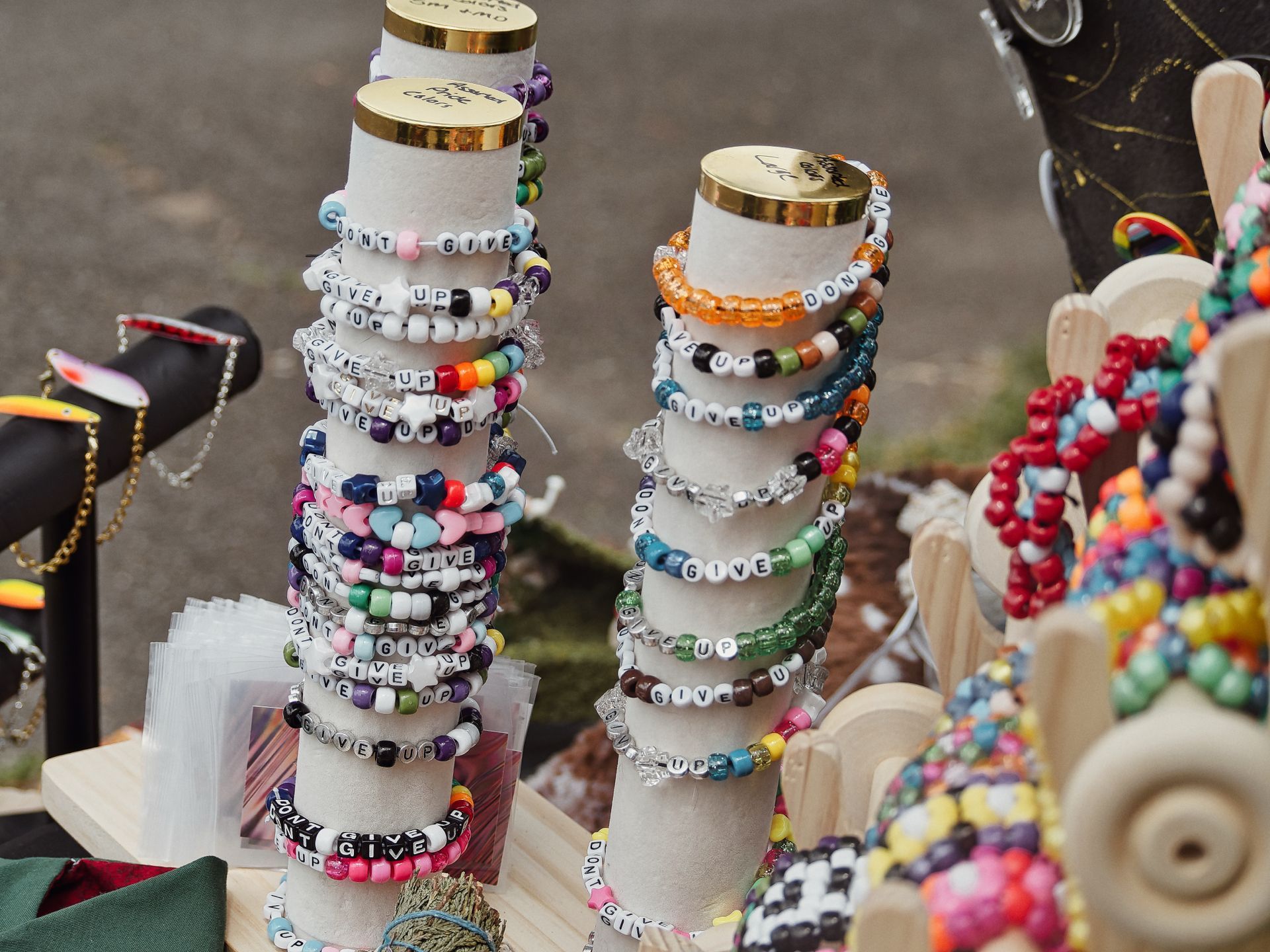 Two foam display cones stacked with dozens of colorful beaded bracelets reading "DON'T GIVE UP" in white letter beads, topped with gold lids, displayed on a wooden tray at what appears to be an outdoor market stall. Multicolored bead figurines on wooden frames crowd the right side, with jewelry displays blurred softly on the left.