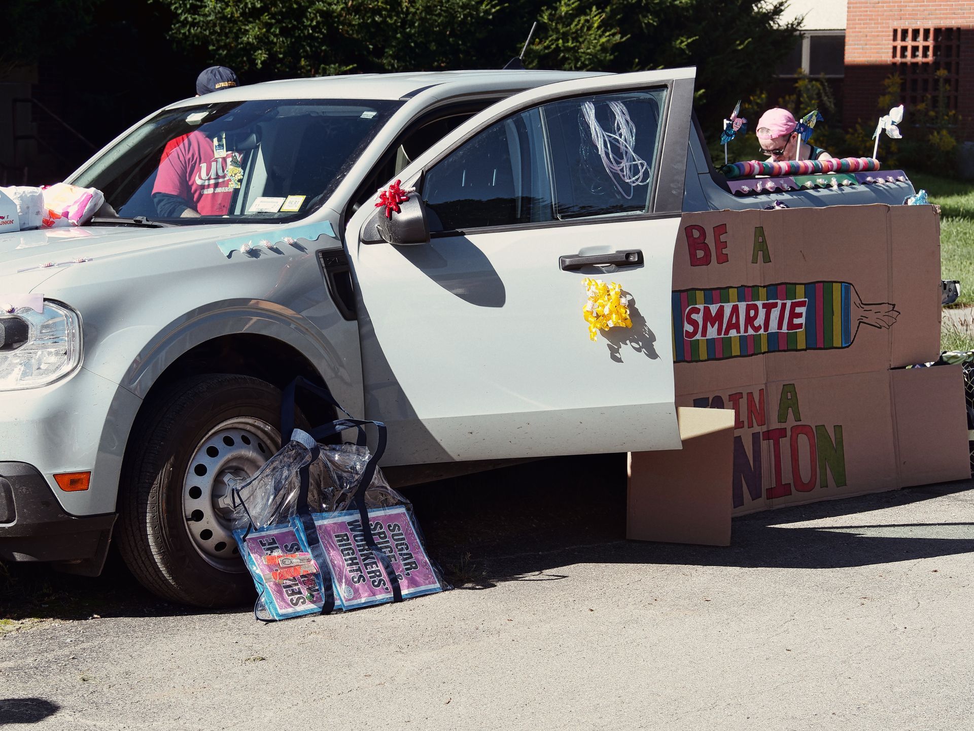 A white compact car decorated with yellow ribbon flowers on the door and red bows on the mirror, parked next to a large cardboard sign reading "BE A SMARTIE / JOIN A UNION" in colorful painted letters. Tote bags leaning against the front tire are labeled "SUGAR," "WORKERS RIGHTS," and partial text, while a person in a pink head wrap stands behind the trunk, and a driver in a red shirt and baseball cap sits inside.