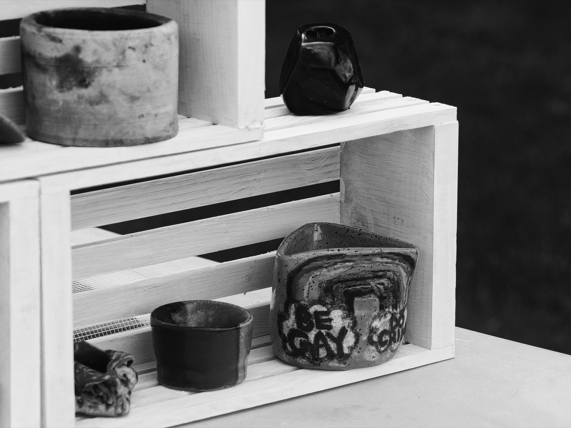 Black-and-white shot of handmade ceramic vessels arranged on and inside a white-painted wooden slatted crate. A weathered cylindrical pot sits on a surface to the upper left, a small dark glazed vessel perches on the crate's top edge, and inside the lower shelf sit a dark ceramic bowl and a rough, irregularly shaped ceramic piece with the words "BE TODAY DO GO" painted in bold lettering across it.