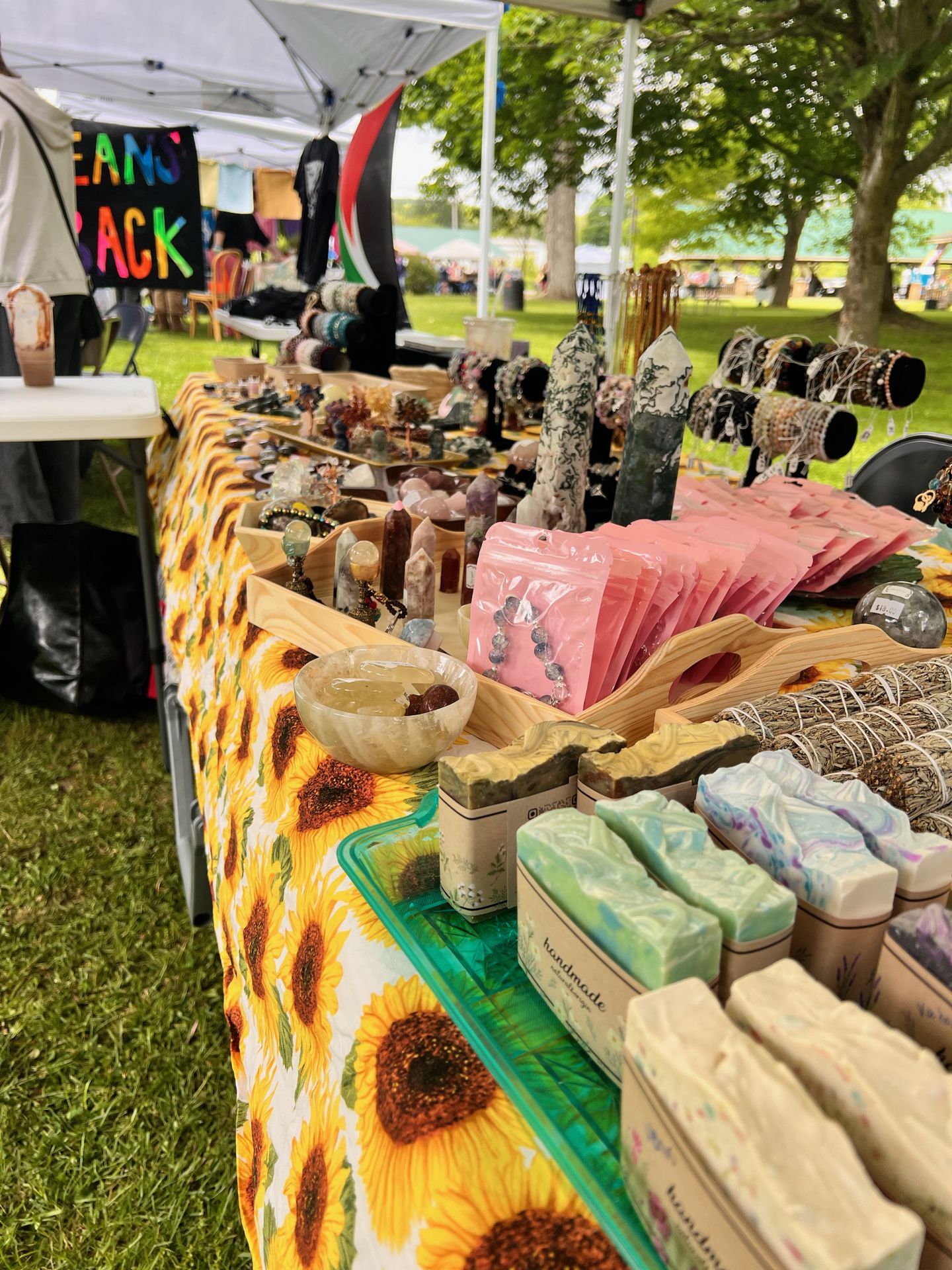 Outdoor craft market table draped in a sunflower-print cloth, loaded with crystals, polished stone towers, beaded jewelry on display stands, pink packaging pouches, a honey-colored alabaster bowl, bundled white sage smudge sticks, and rows of swirled handmade soap bars labeled "handmade" in the foreground. A white pop-up canopy overhead and lush green trees blur into the background, with a colorful banner on the left partially reading "EAMS ACK."