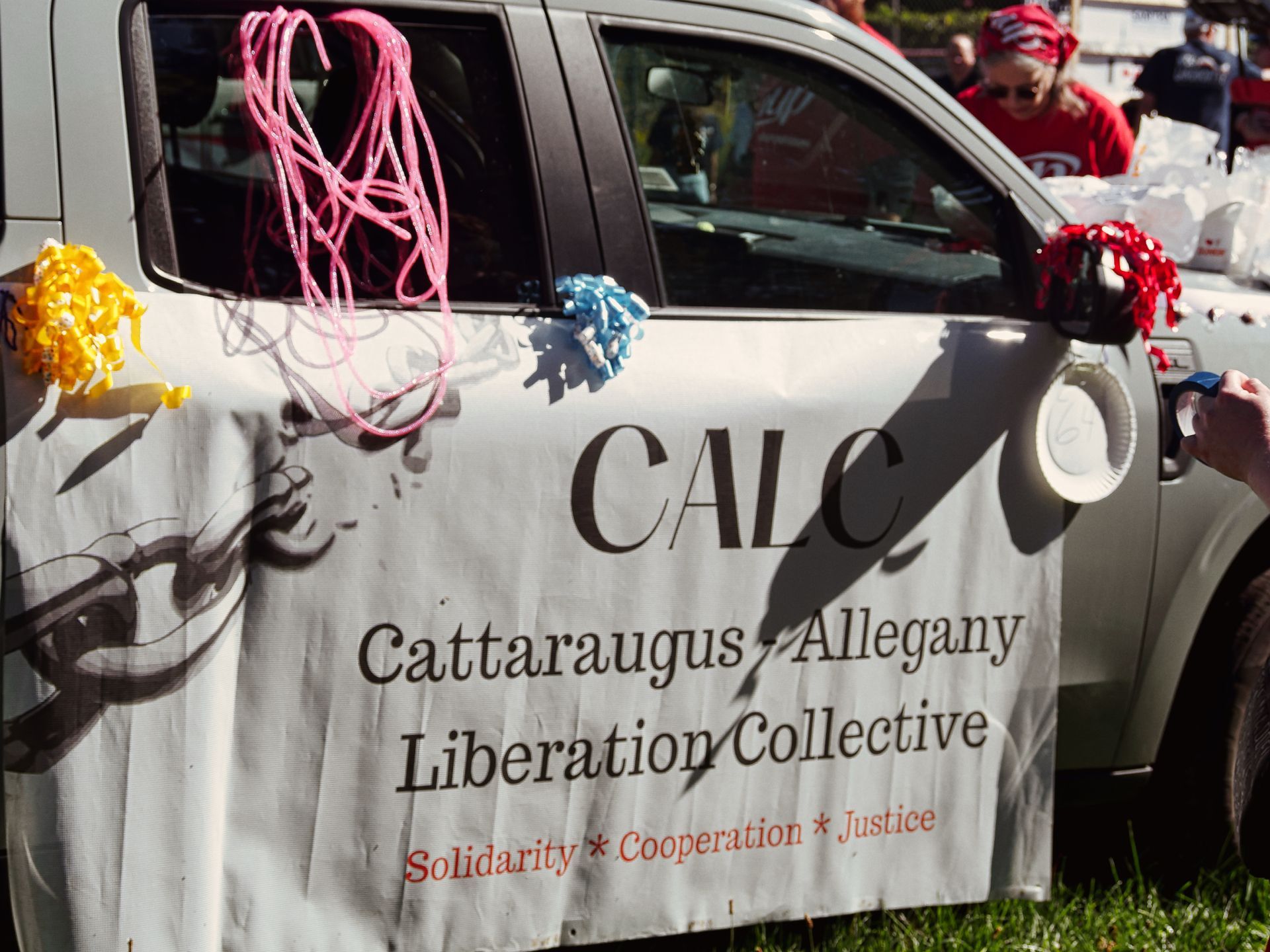 A white pickup truck decorated with colorful ribbon pompoms and looped yarn in pink, yellow, blue, and red, bearing a banner that reads "CALC / Cattaraugus Allegany Liberation Collective / Solidarity * Cooperation * Justice." In the background, participants in red shirts handle white bags, blurred in the shallow depth of the background.
